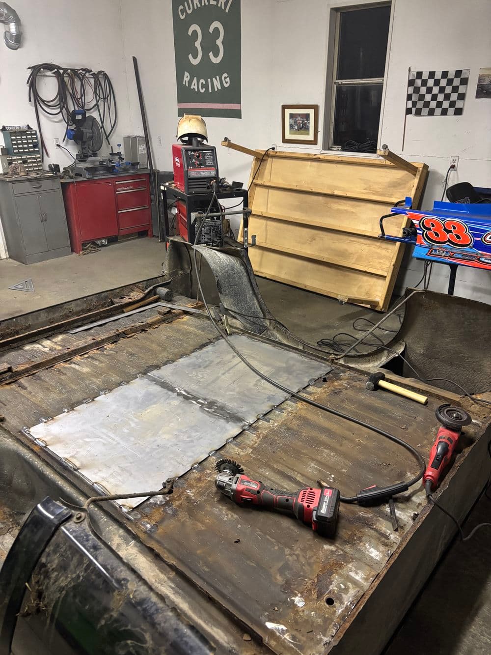 Race car workshop with metal flooring repair in progress, tools and race memorabilia visible.